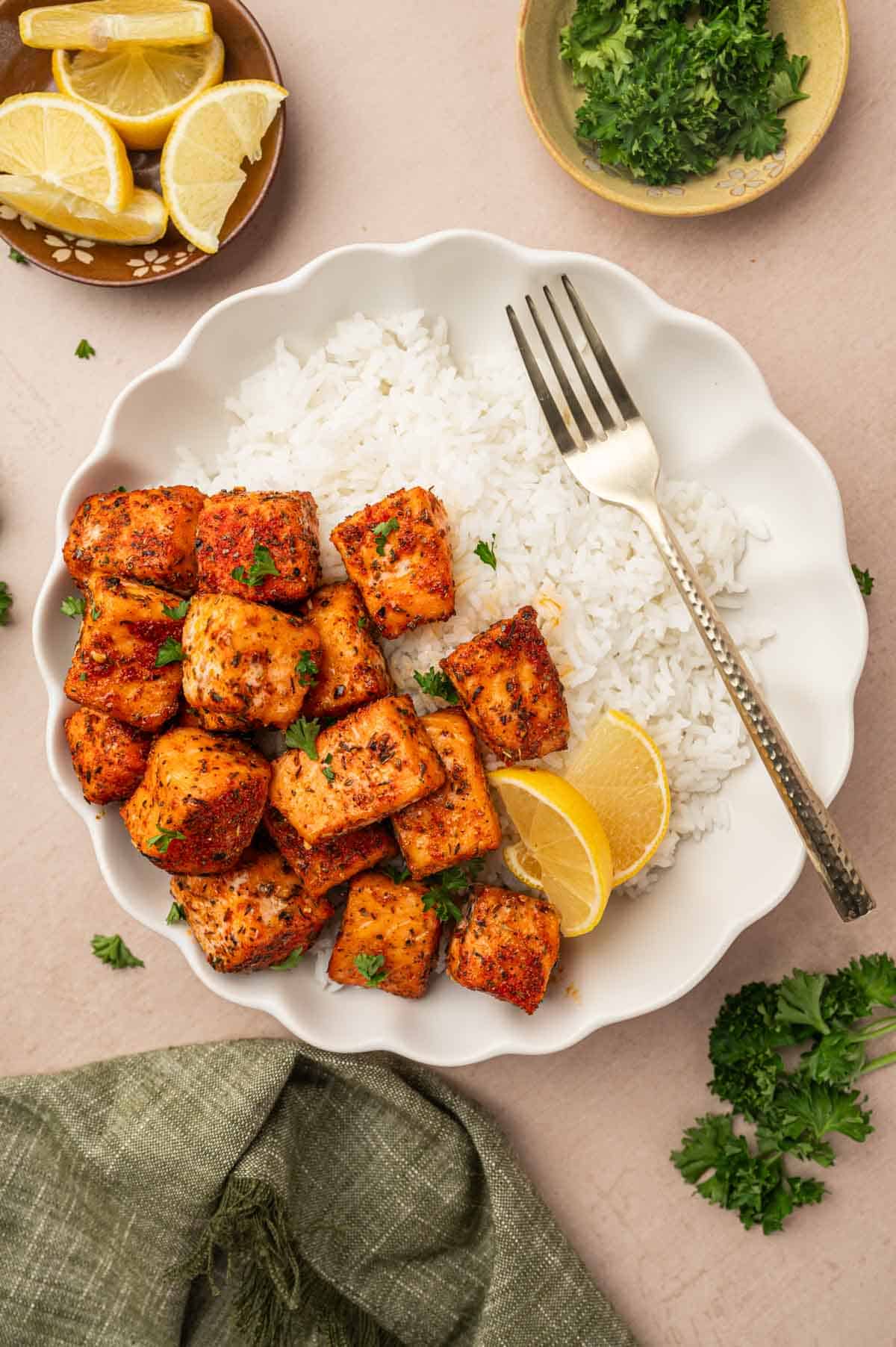 In a white bowl the blackened salmon bites served over white rice, garnish with parsley and lemon wedges.