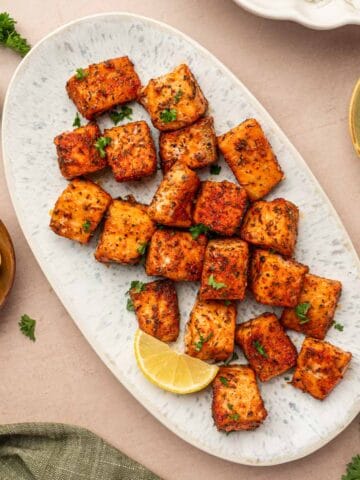 Blackened salmon bites in a oval platter arranged beautifully and garnished with parsley and lemon wedges in the side.