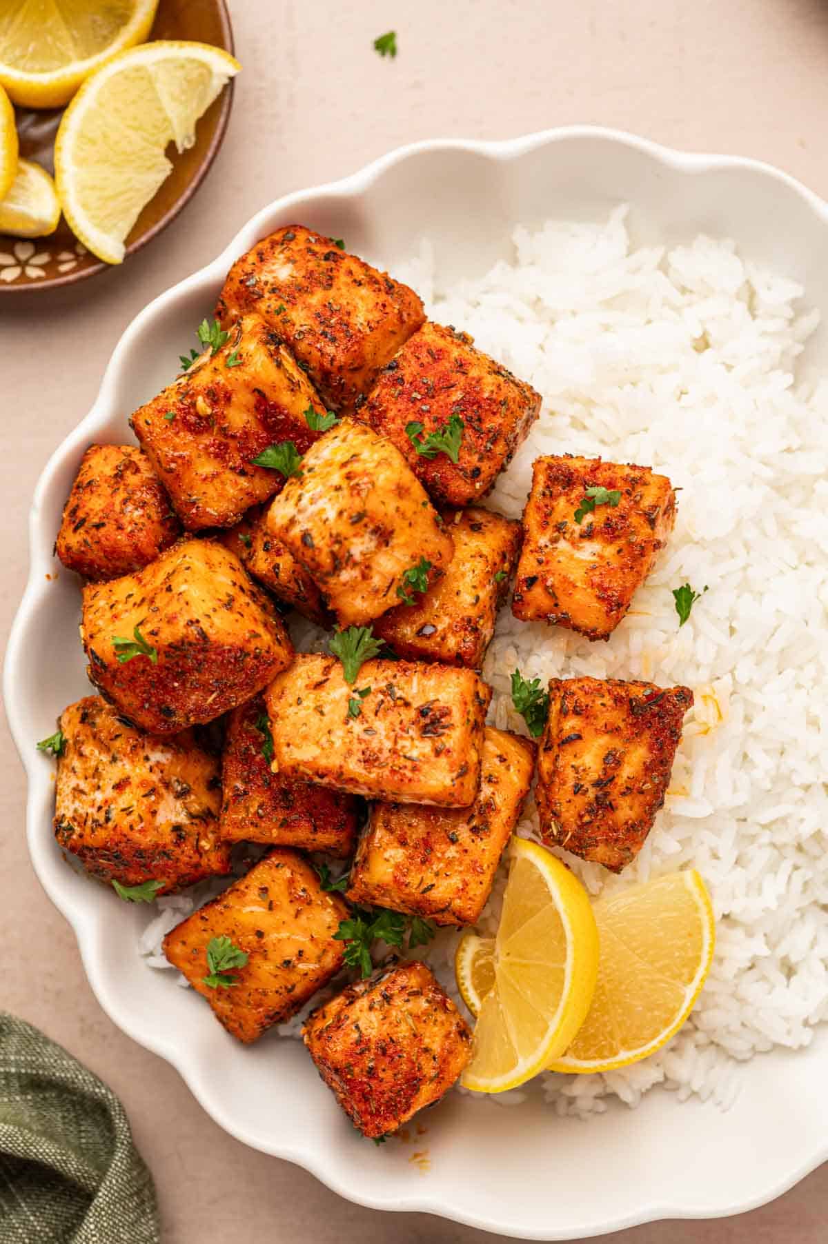 Blackened salmon bites in a bowl with white rice, ganrished with parsley and served with lemon wedges on the sides.