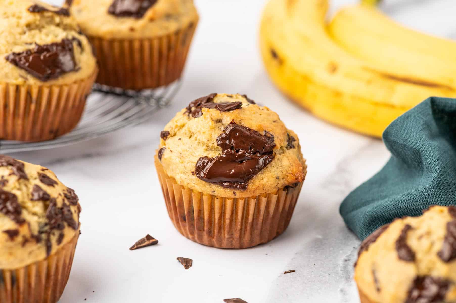 Horizontal image of banana chocolate chunk muffins placed beautifully on a marble surface, banana's in the background.