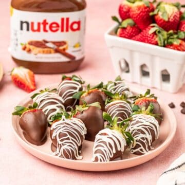 Nutella covered strawberries in a pink plate with Nutella jar and fresh strawberries behind.