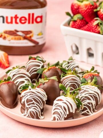 Nutella covered strawberries in a pink plate with Nutella jar and fresh strawberries behind.