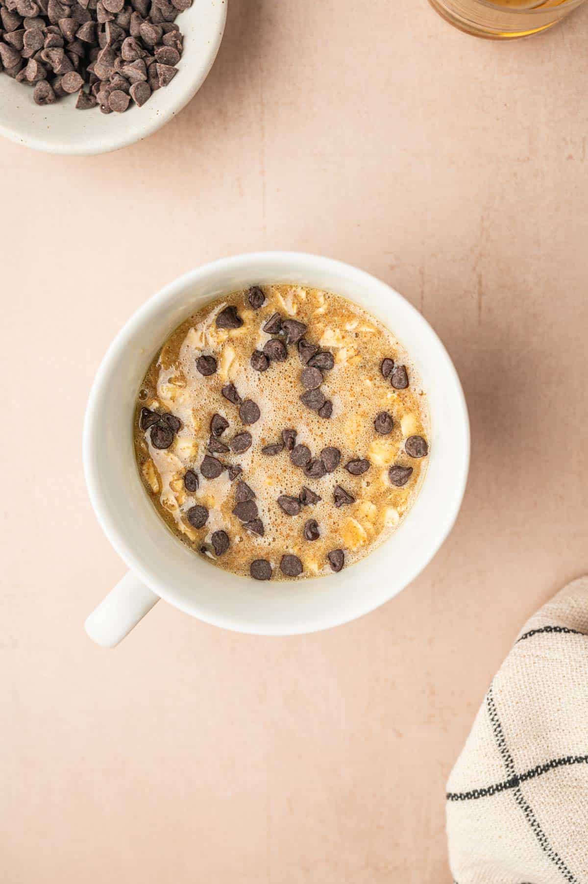 Mini chocolate chips added to the oatmeal mug cake batter in white mug.