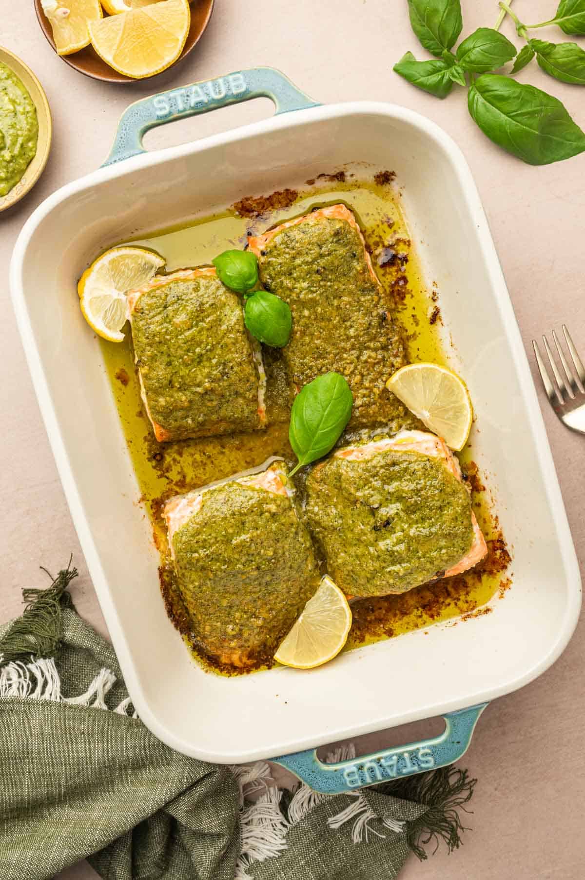 Baked Pesto butter salmon in a baking dish. Garnished with basil. Lemon wedges in a bowl aside.