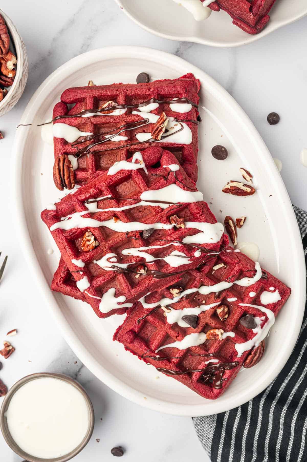 Red velvet cake waffles in a oval plate. Dark chocolate ganche and cream cheese drizzle topped with chopped pecans and chocolate chips.