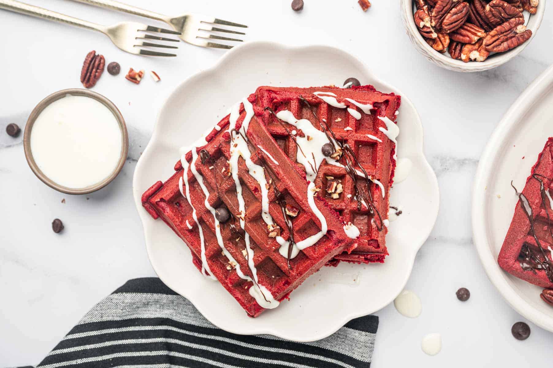 In a white plate, red velvet cake mix waffles topped with pecans and chocolate chips with cream cheese drizzle and Chocolate ganache.