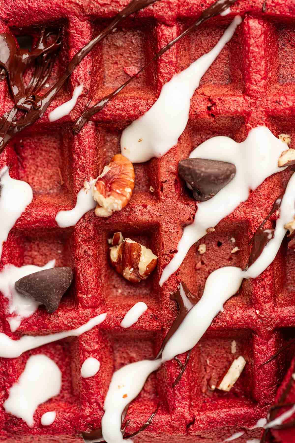 Close up shot of red velvet cake mix waffles topped with pecans and chocolate chips with cream cheese drizzle and Chocolate ganache.