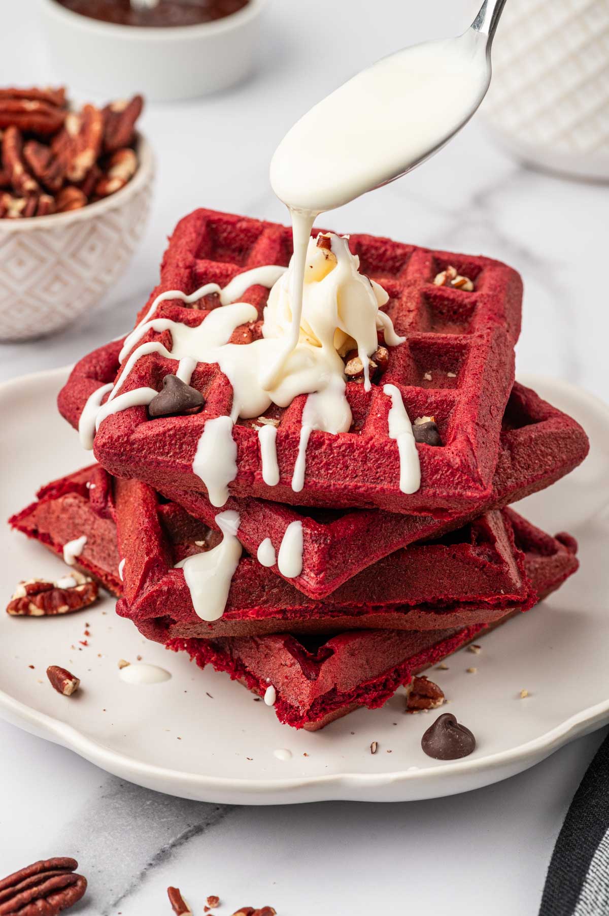 Cream cheese drizzle over the stacked red velvet cake waffles in a plate and pecans in the behind.