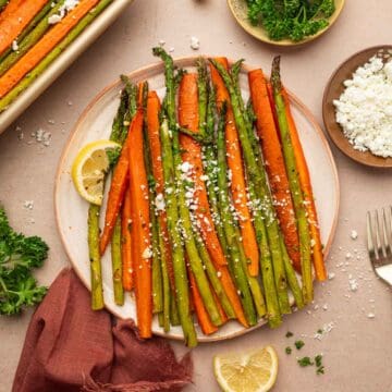 Roasted carrots and asparagus with goat cheese plated in a off white plate served with lemon wedges in the sides and garnished with parsley.