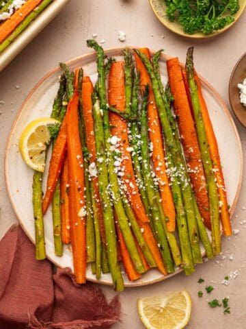 Roasted carrots and asparagus with goat cheese plated in a off white plate served with lemon wedges in the sides and garnished with parsley.