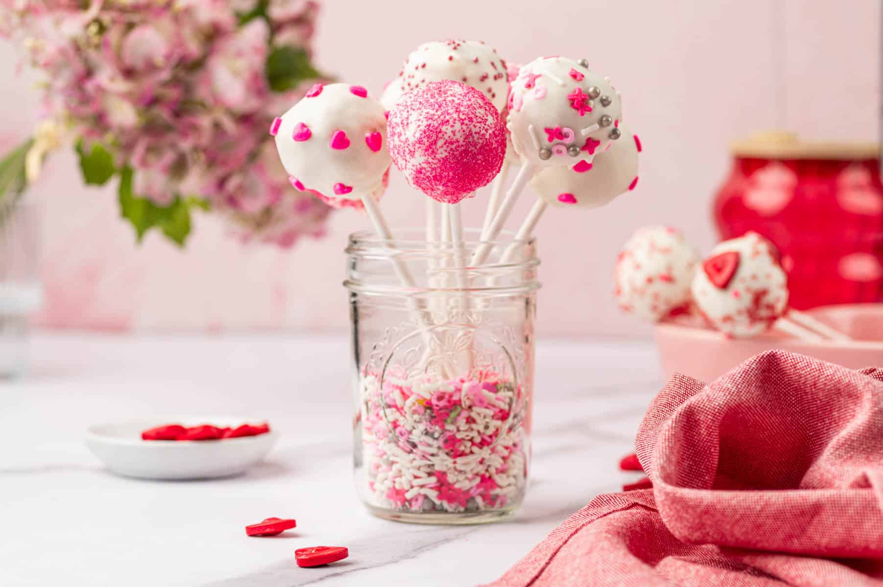 Horizontal Image of Valentines cake pops arranged in a glass jar with a pink linens on the saide.