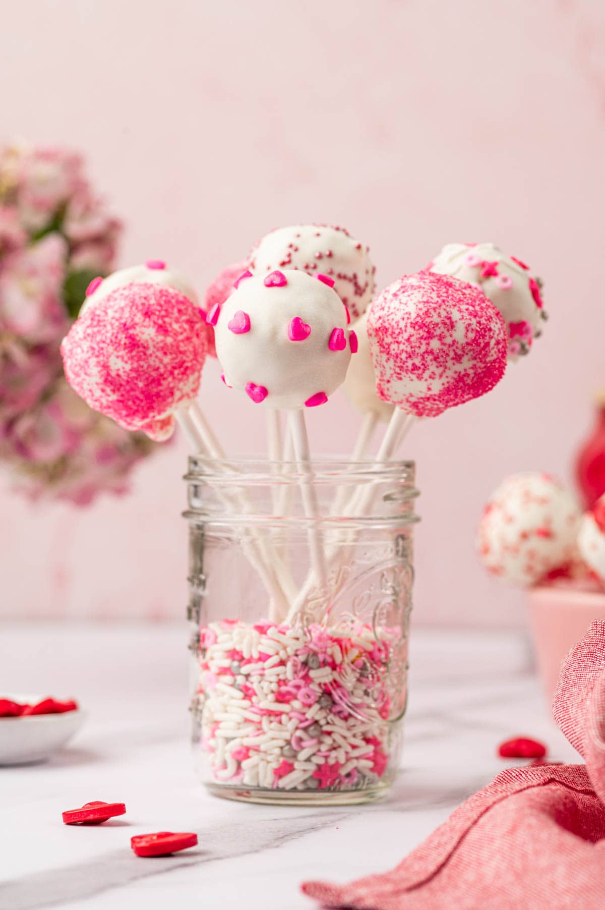 Valentine cake pops arranged in a glass jar filled with sprinkles.