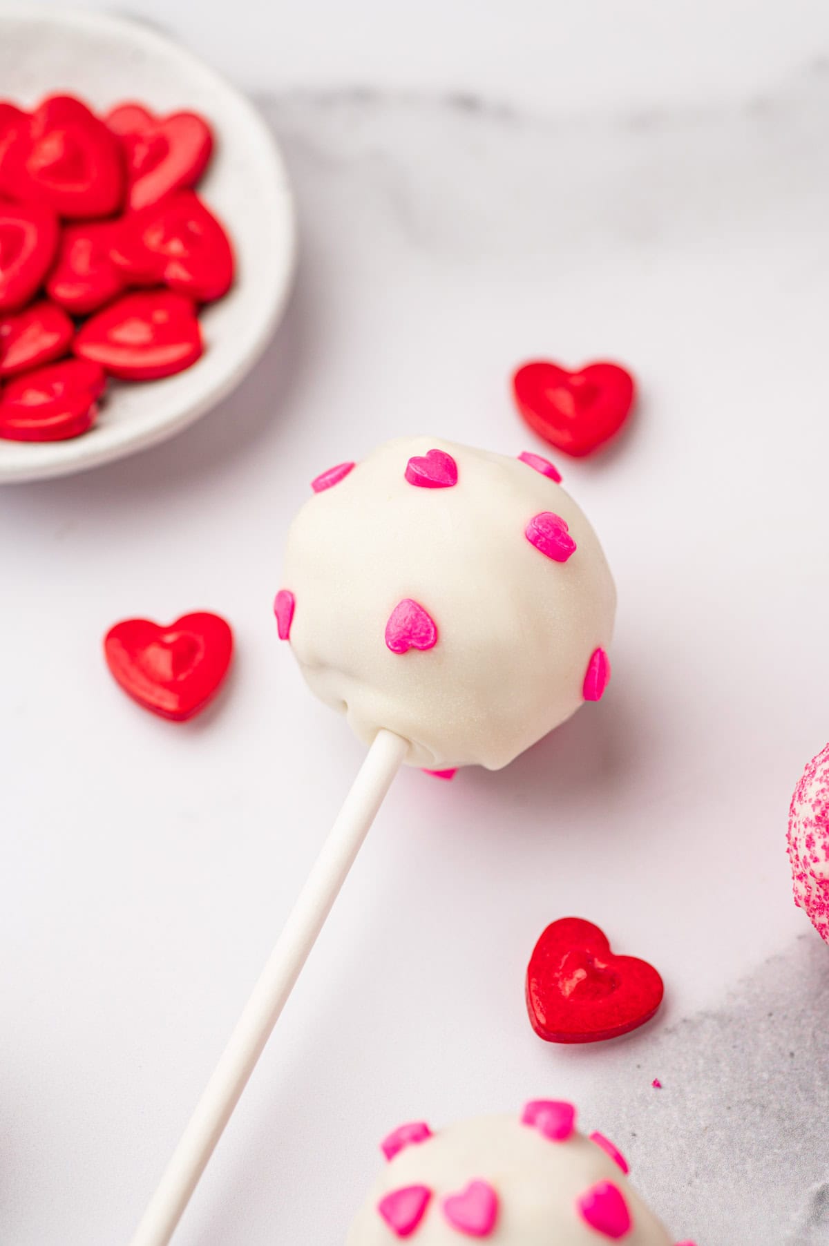 Pink heart decorated valentine cake pops placed in a marble table with red heart sprinkles in the side.