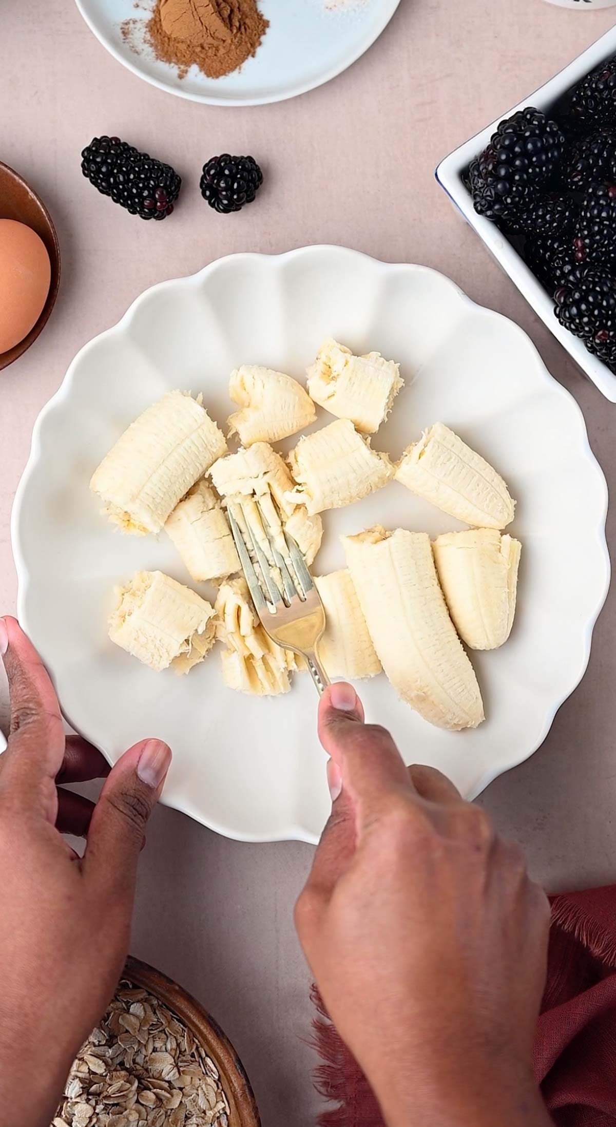 A hand is mashing the banana with a fork in a white bowl for making the muffins.