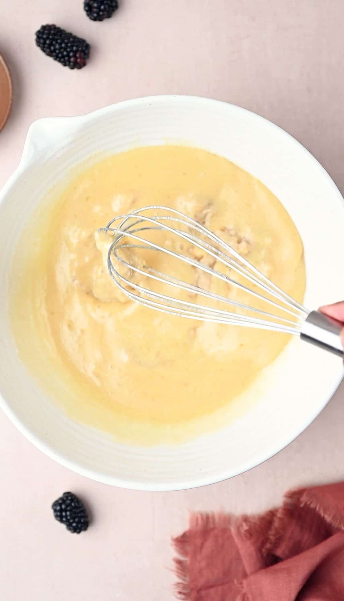 In a large white bowl the wet ingredients are being mixed with a whisk for making Banana Blackberry Oatmeal Muffins.