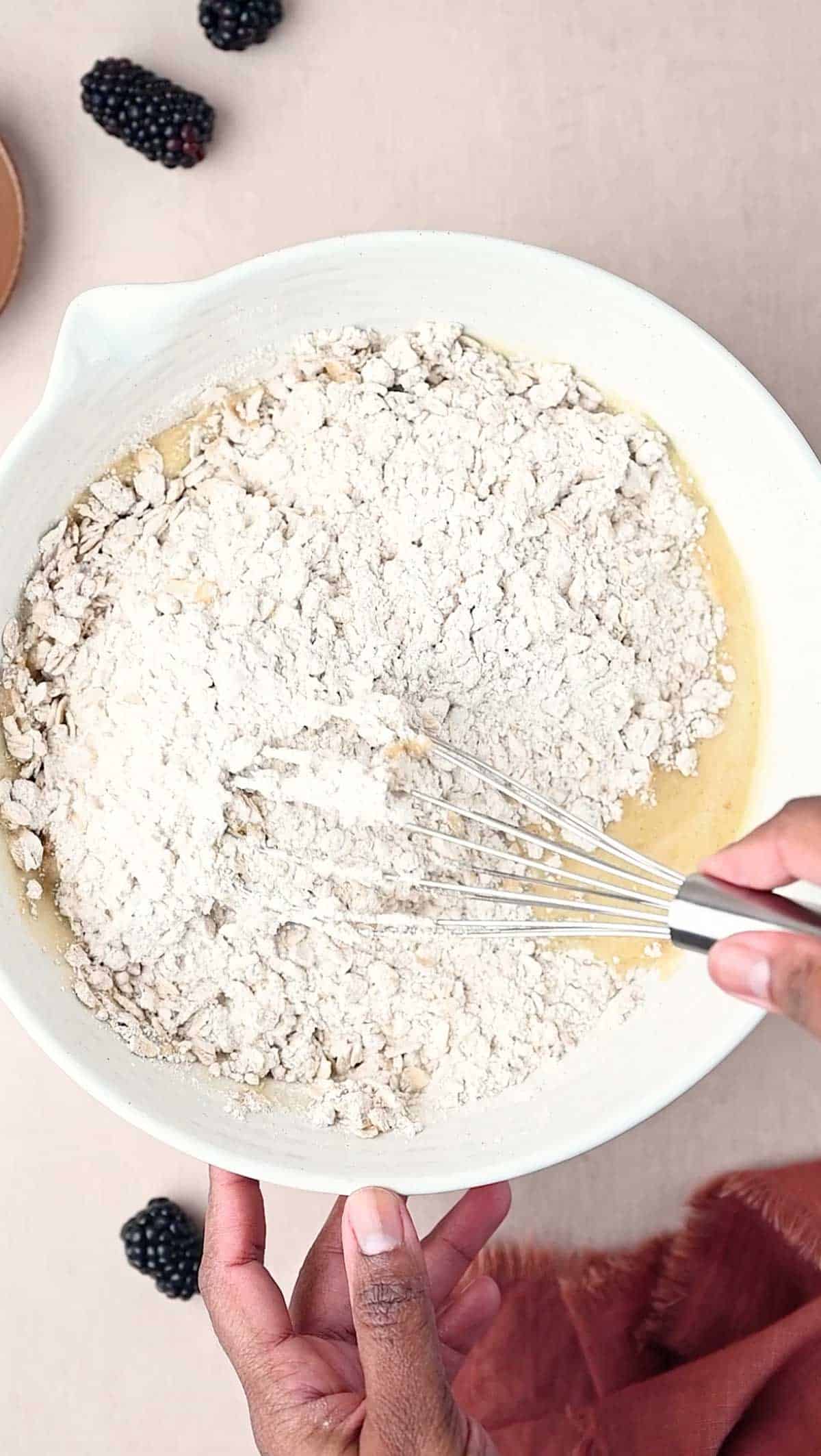 The wet and dry ingredients being mixed in white bowl for Banana Blackberry Oatmeal Muffins recipe.