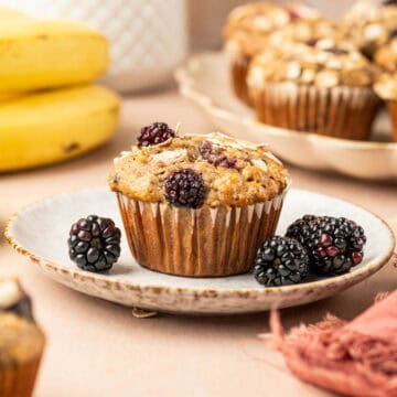 Square image of Single banana blackberry oatmeal muffin placed in a plate with fresh blackberries arround and banana is the behind.