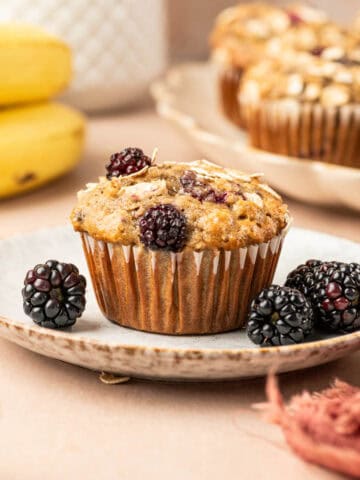 Square image of Single banana blackberry oatmeal muffin placed in a plate with fresh blackberries arround and banana is the behind.