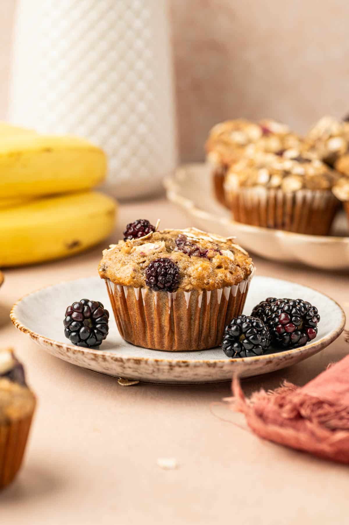 Single banana blackberry oatmeal muffin placed in a plate with fresh blackberries arround and banana is the behind.