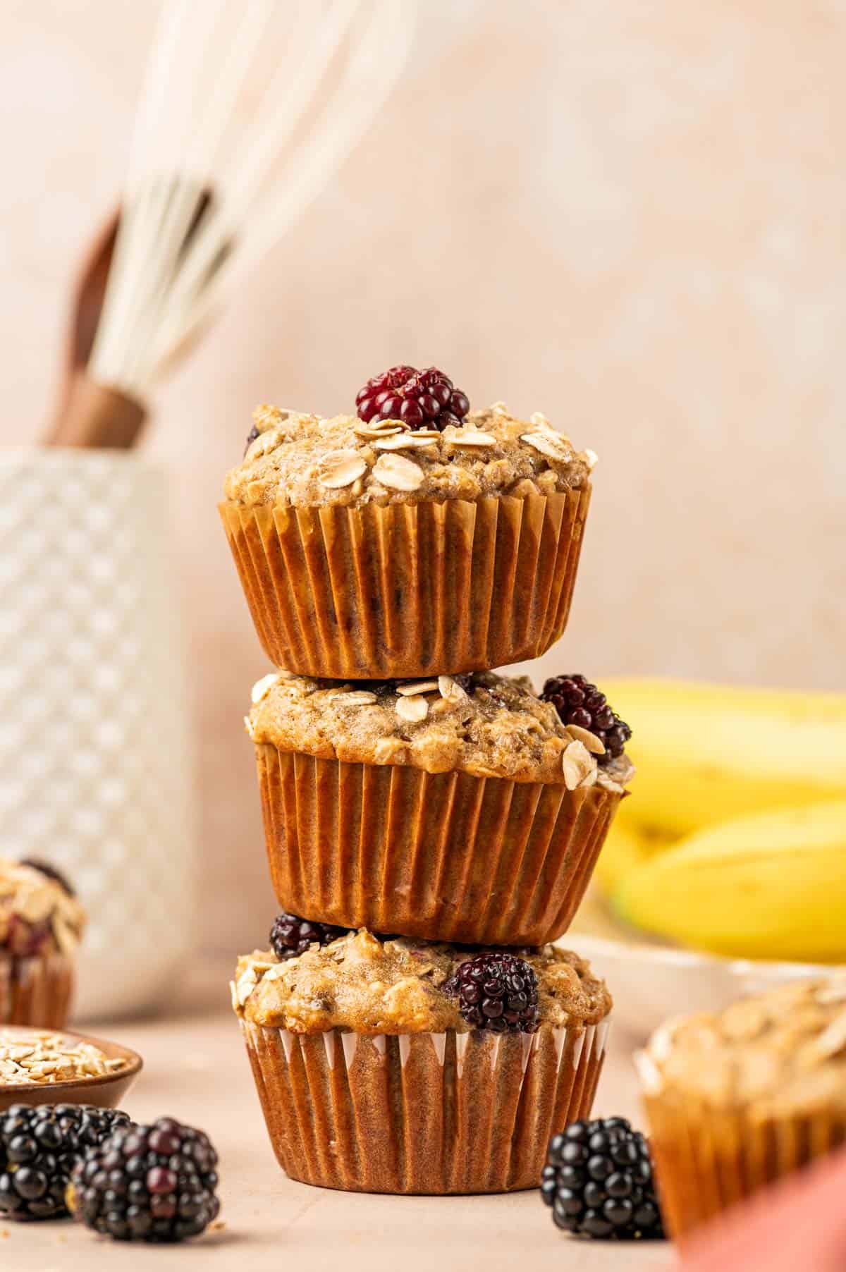 Three Banana blackberry oatmeal muffins stacked with fresh blackberries arround and banana is the behind.