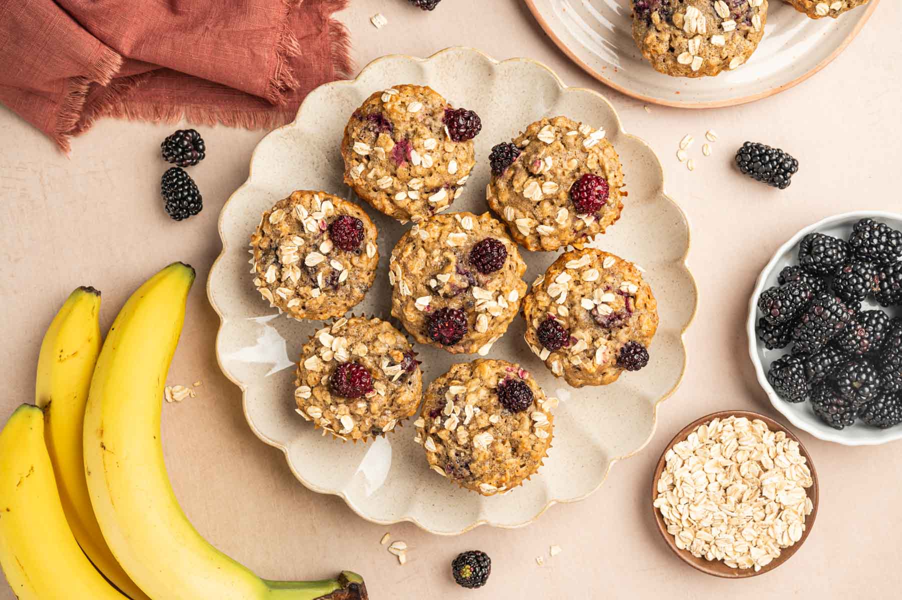 Horizontal Image of banana blackberry oatmeal muffin placed in a plate with fresh blackberries arround and banana is the behind.