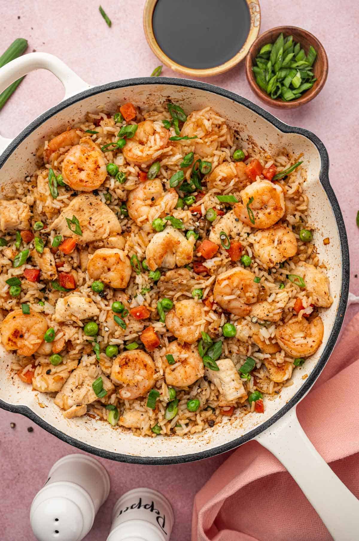 Cooked Chicken and shrimp fried rice in a white skillet surrounded by a wooden bowl of green onions, soy sauce in a bowl, salt and pepper shakes in the bottom.