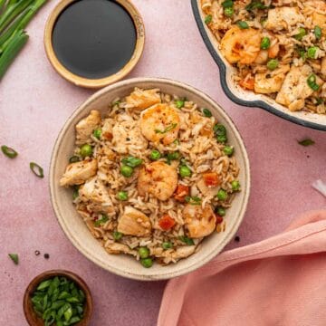 Chicken and shrimp fried rice is placed in a serving bowl, the skillet the fried rice is peeking. Soy sauce is placed in a dipping saucer along with the green onions in a wooden bowl.