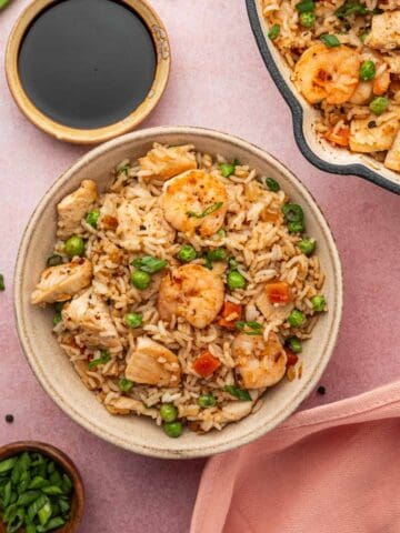 Chicken and shrimp fried rice is placed in a serving bowl, the skillet the fried rice is peeking. Soy sauce is placed in a dipping saucer along with the green onions in a wooden bowl.