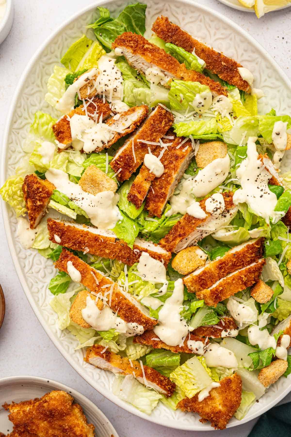 Crispy Chicken caesar salad served in a white oval platter and caesar dressing is drizzled over.