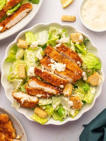 Crispy chicken caesar salad in a white bowl accompanied by a bowl homemade caesar salad dressing.