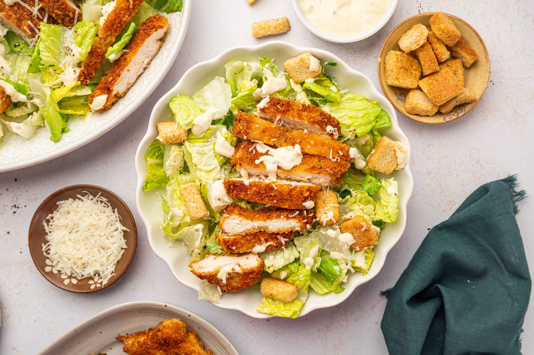 Crispy chicken caesar salad in a white bowl accompanied by a bowl homemade caesar salad dressing with parmesan cheese in a bowl on other side.