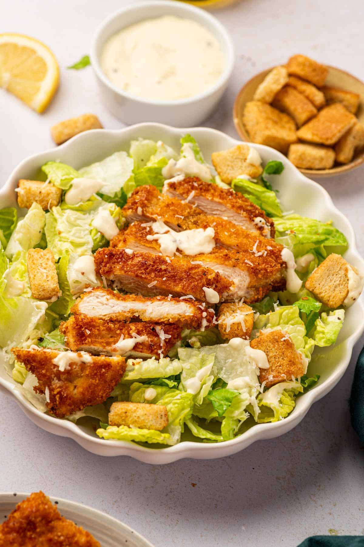 Black light shot of Crispy chicken caesar salad in a white bowl accompanied by a bowl homemade caesar salad dressing.