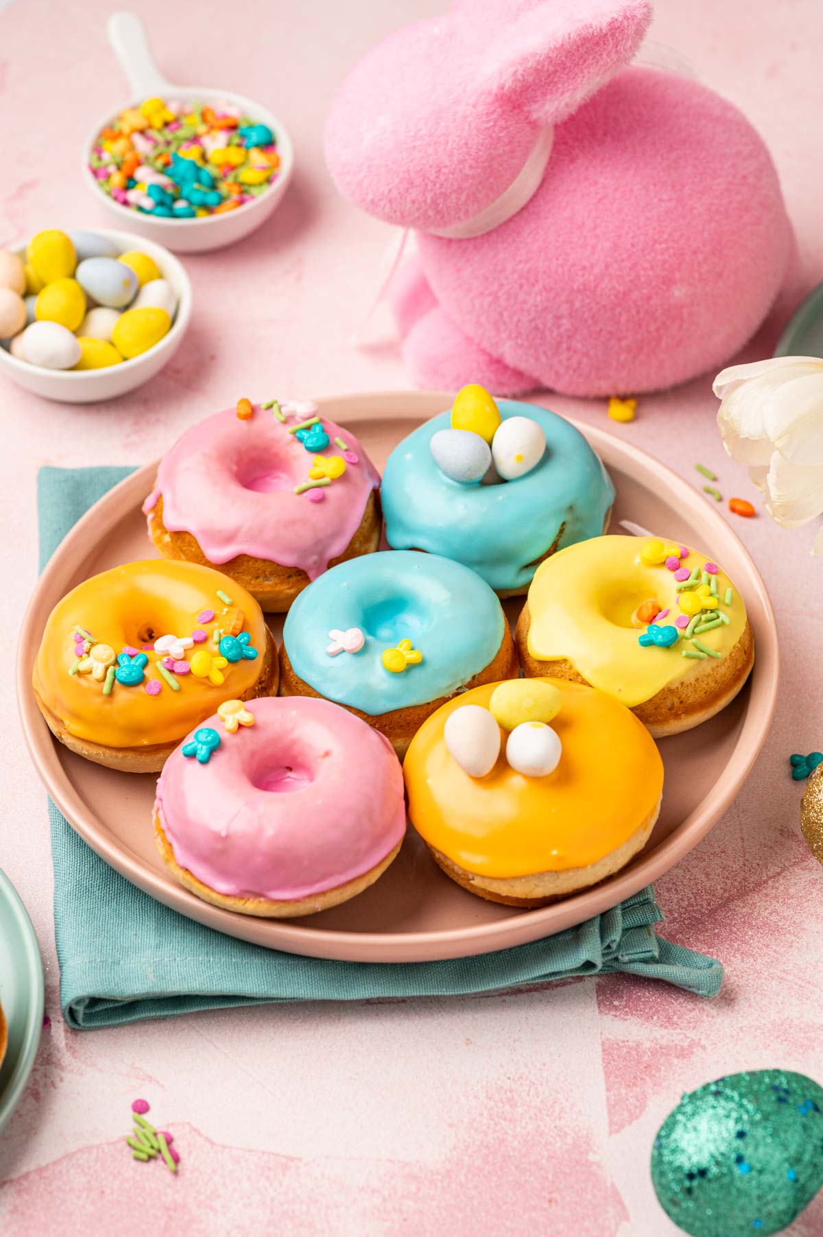 Beautifully decorated homeade baked easter donuts with patel colored donut glaze topped with Easter festive sprinkles, mini eggs. The Easter are placed in a pink plate with blue napkin underneath on a pink backgorund with festive sprinkles aorund.