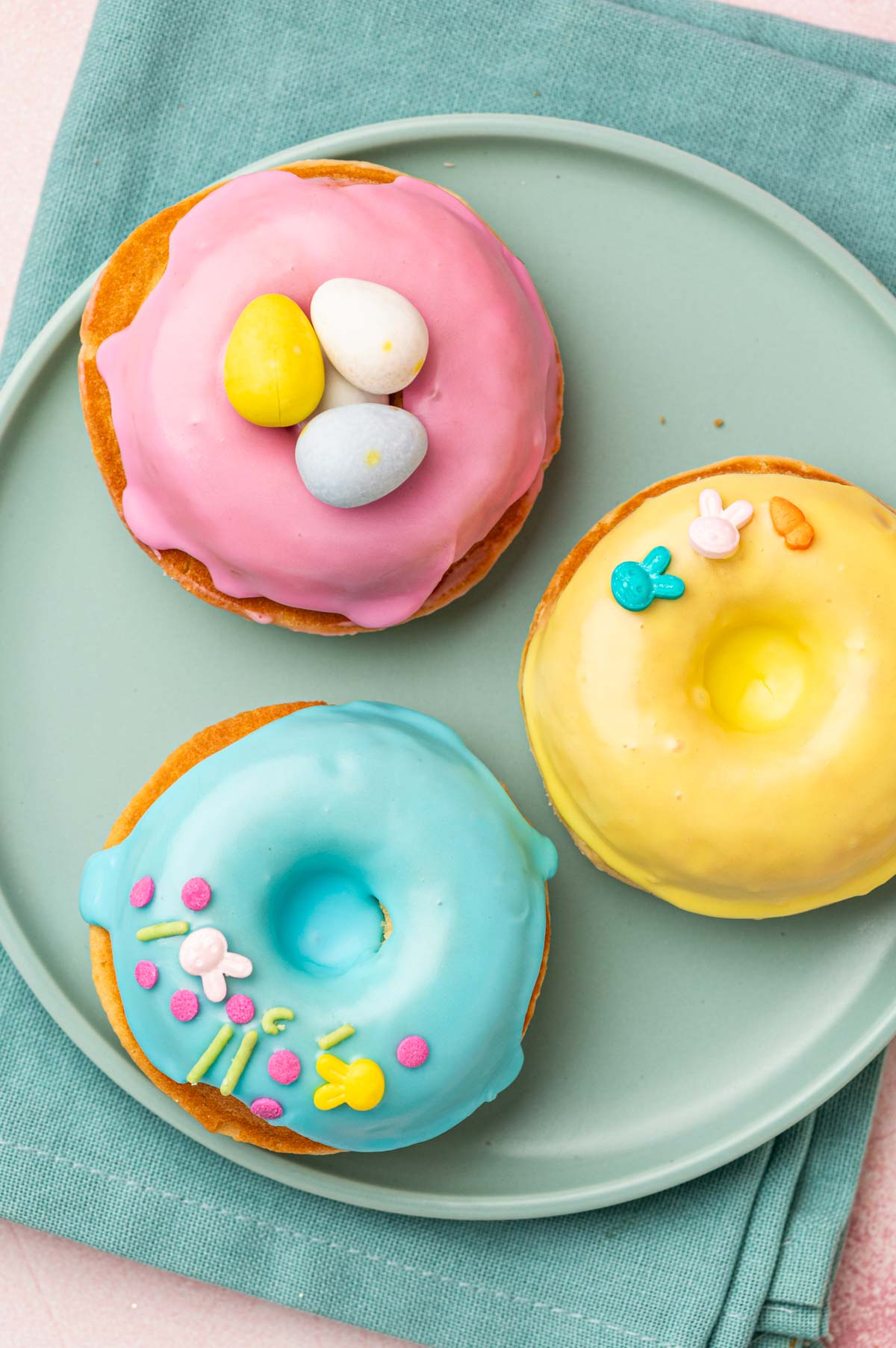 Beautifully decorated homeade baked easter donuts with patel colored donut glaze topped with Easter festive sprinkles, mini eggs. The Easter are placed in a blue plate with blue napkin underneath on a pink backgorund.