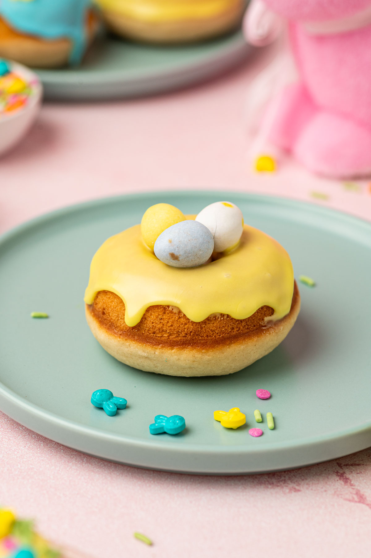 Beautifully decorated single easter donuts with pastel colored donut glaze topped with Easter festive sprinkles, mini eggs. The Easter are placed in a blue plate on a pink backgorund with festive sprinkles, mini eggs around.