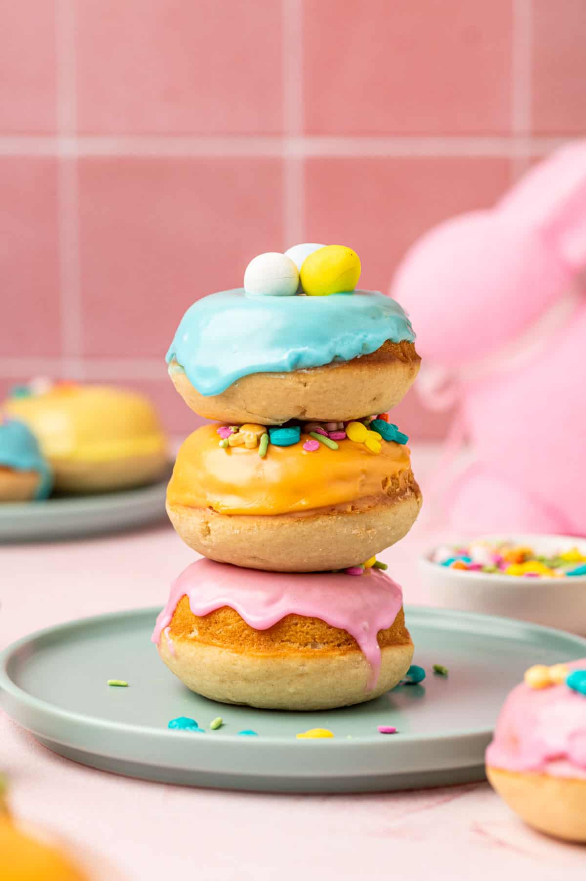 Stacked baked easter donuts one above the other in a pink background on a blue plate. easter donuts with pastel colored donut glaze topped with Easter festive sprinkles, mini eggs. 