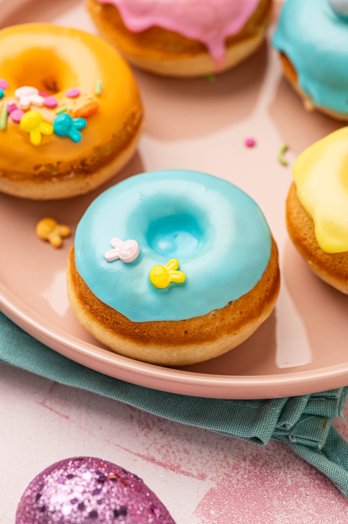 CLose up shot of beautifully decorated homeade baked easter donuts with pastel colored donut glaze topped with Easter festive sprinkles, mini eggs. The Easter are placed in a pink plate with blue napkin underneath on a pink backgorund with festive sprinkles, mini eggs around.