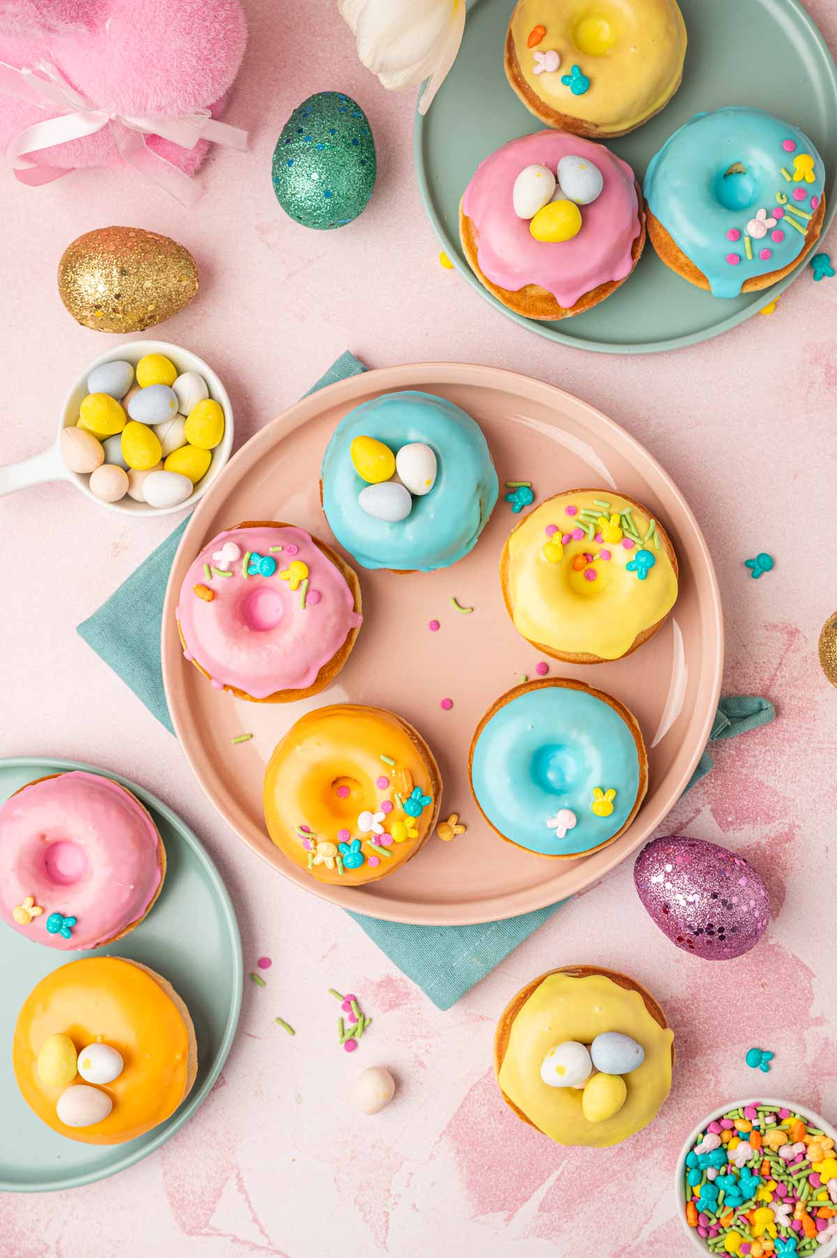 Beautifully decorated homeade baked easter donuts with pastel colored donut glaze topped with Easter festive sprinkles, mini eggs. The Easter are placed in a pink and blue plates with blue napkin underneath on a pink backgorund with festive sprinkles, mini eggs around.