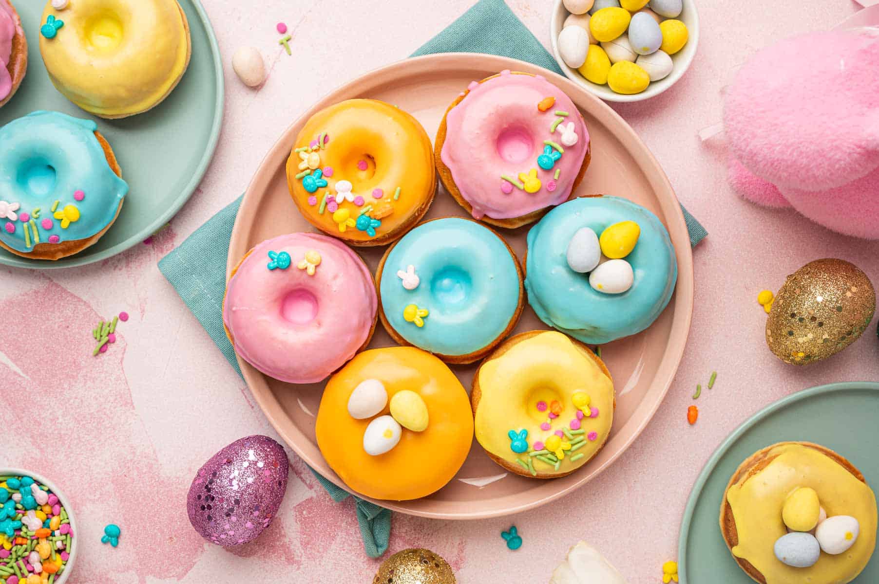 Horizontal image of Beautifully decorated homeade baked easter donuts with pastel colored donut glaze topped with Easter festive sprinkles, mini eggs. The Easter are placed in a pink and blue plates with blue napkin underneath on a pink backgorund with festive sprinkles, mini eggs around.