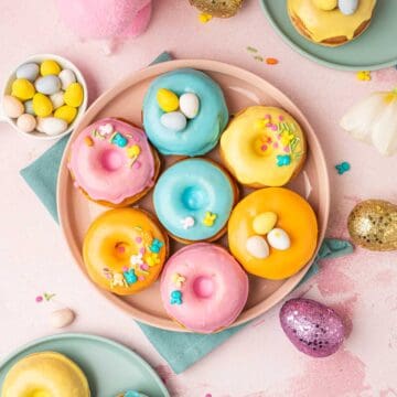 Beautifully decorated homeade baked easter donuts with patel colored donut glaze topped with Easter festive sprinkles, mini eggs. The Easter are placed in a pink plate with blue napkin underneath on a pink backgorund with festive sprinkles, mini eggs around.