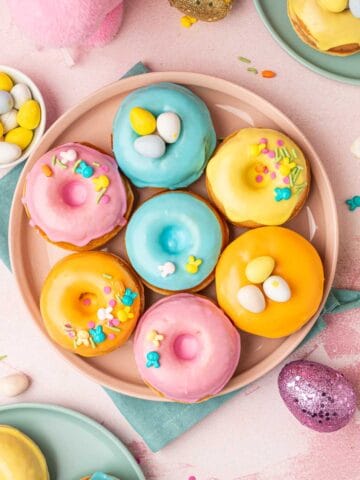 Beautifully decorated homeade baked easter donuts with patel colored donut glaze topped with Easter festive sprinkles, mini eggs. The Easter are placed in a pink plate with blue napkin underneath on a pink backgorund with festive sprinkles, mini eggs around.