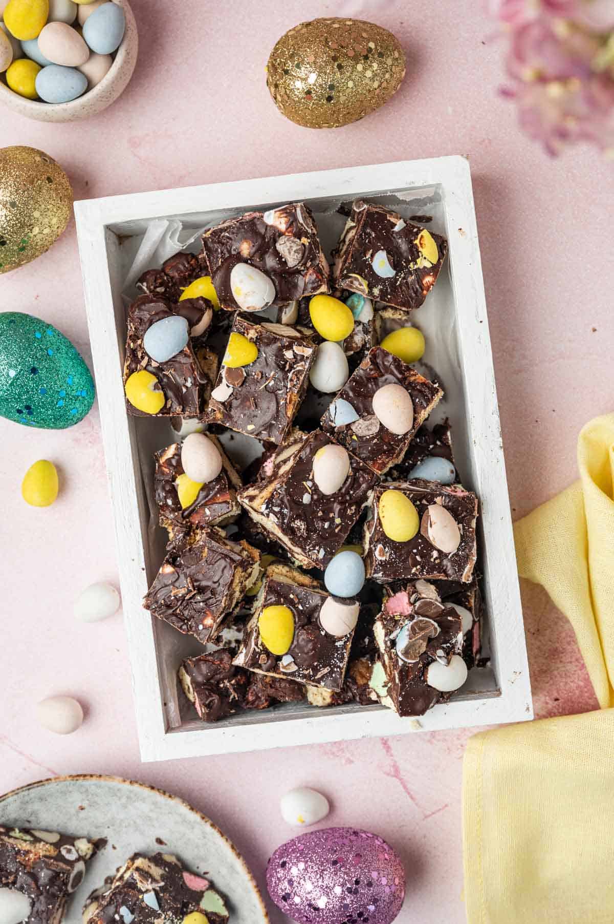 Beautifully decorated Easter Rocky Road fudge placed in a white tray sourranded  by Easter decorations in a pink background.