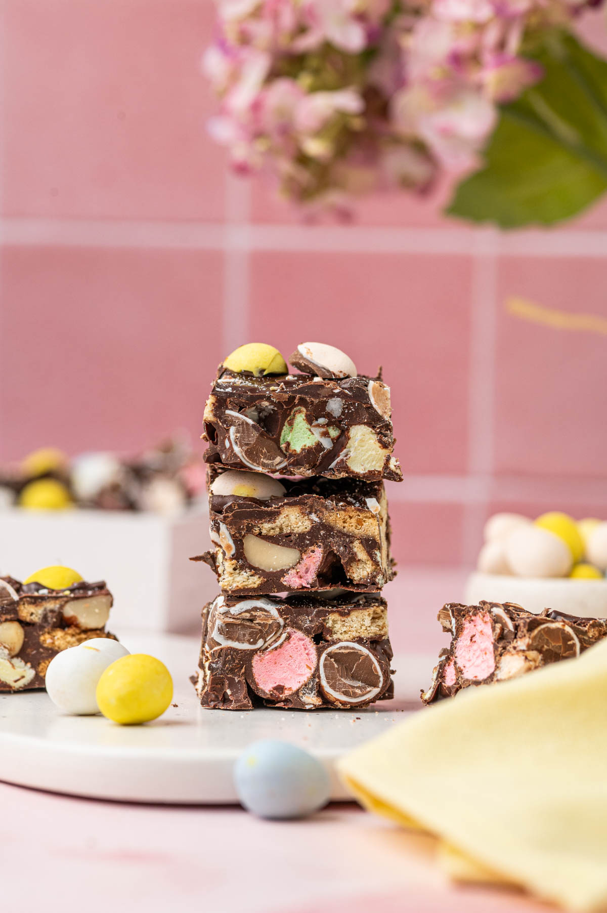 Rocky Road with easter themed fudge stacked one above the other beautifully loaded with mini marshmallows and mini eggs chocolate. With pink flowers behind.