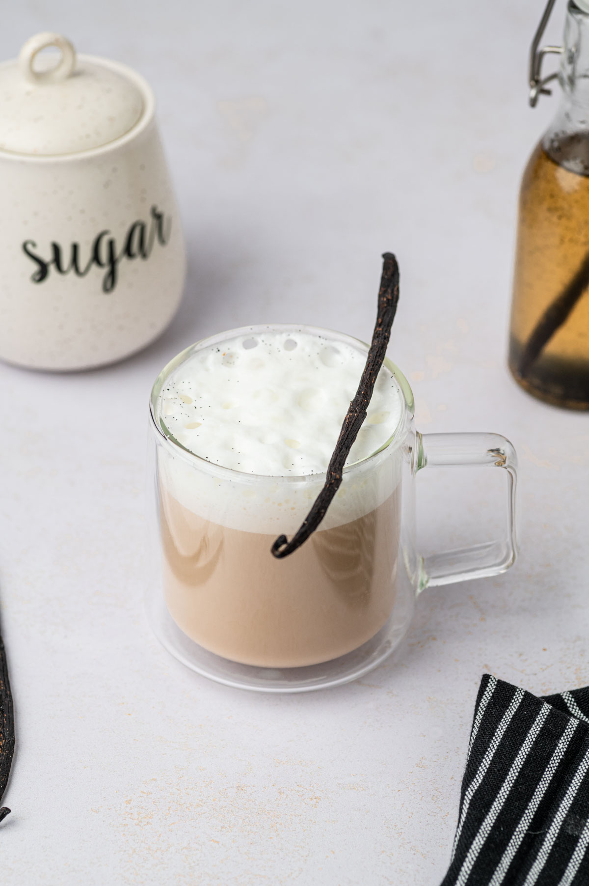 Best vanilla bean latte in a glass mug, vanilla bean on it. In background vanilla bean syrup in a glass jar.