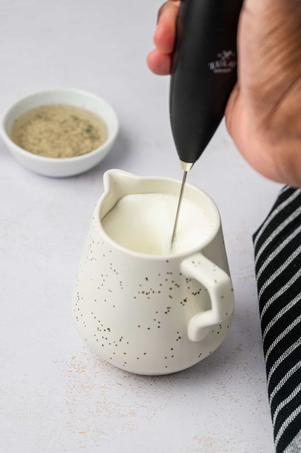 Milk frother being used to make vanilla bean latte recipe in a white creamer mug.