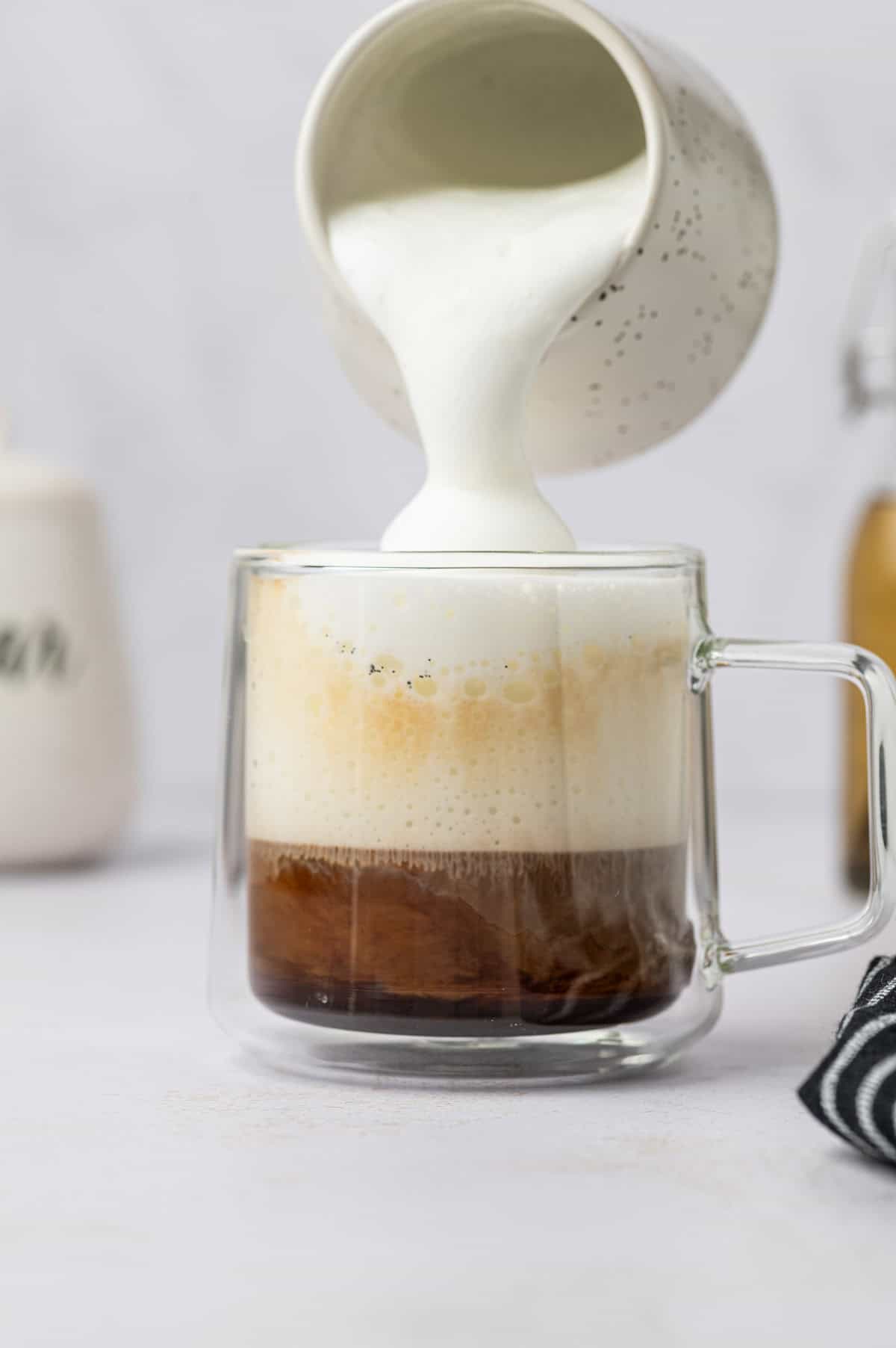 Frothed milk being added to a glass milk with espresso already on it.