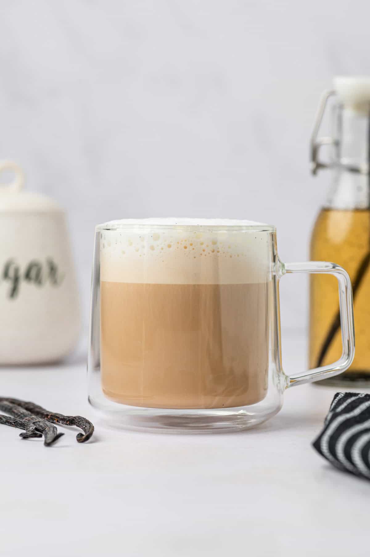 Homemade vanilla bean latte in a glass mug. The vanilla beans are in the side and a homemade vanilla bean coffee syrup on the background.