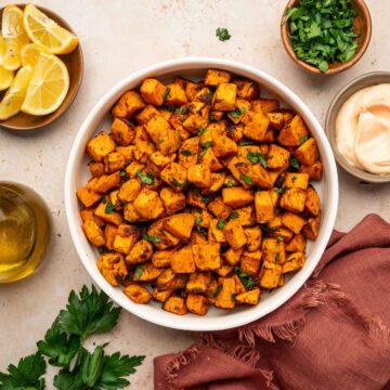 Perfectly crispy sweet potato cubes sprinkled with parsley and sea salt in a white bowl along with dipping sauce, lemon wedges in the sides.