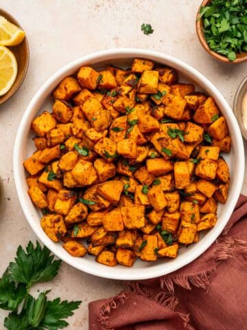 Perfectly crispy sweet potato cubes sprinkled with parsley and sea salt in a white bowl along with dipping sauce, lemon wedges in the sides.