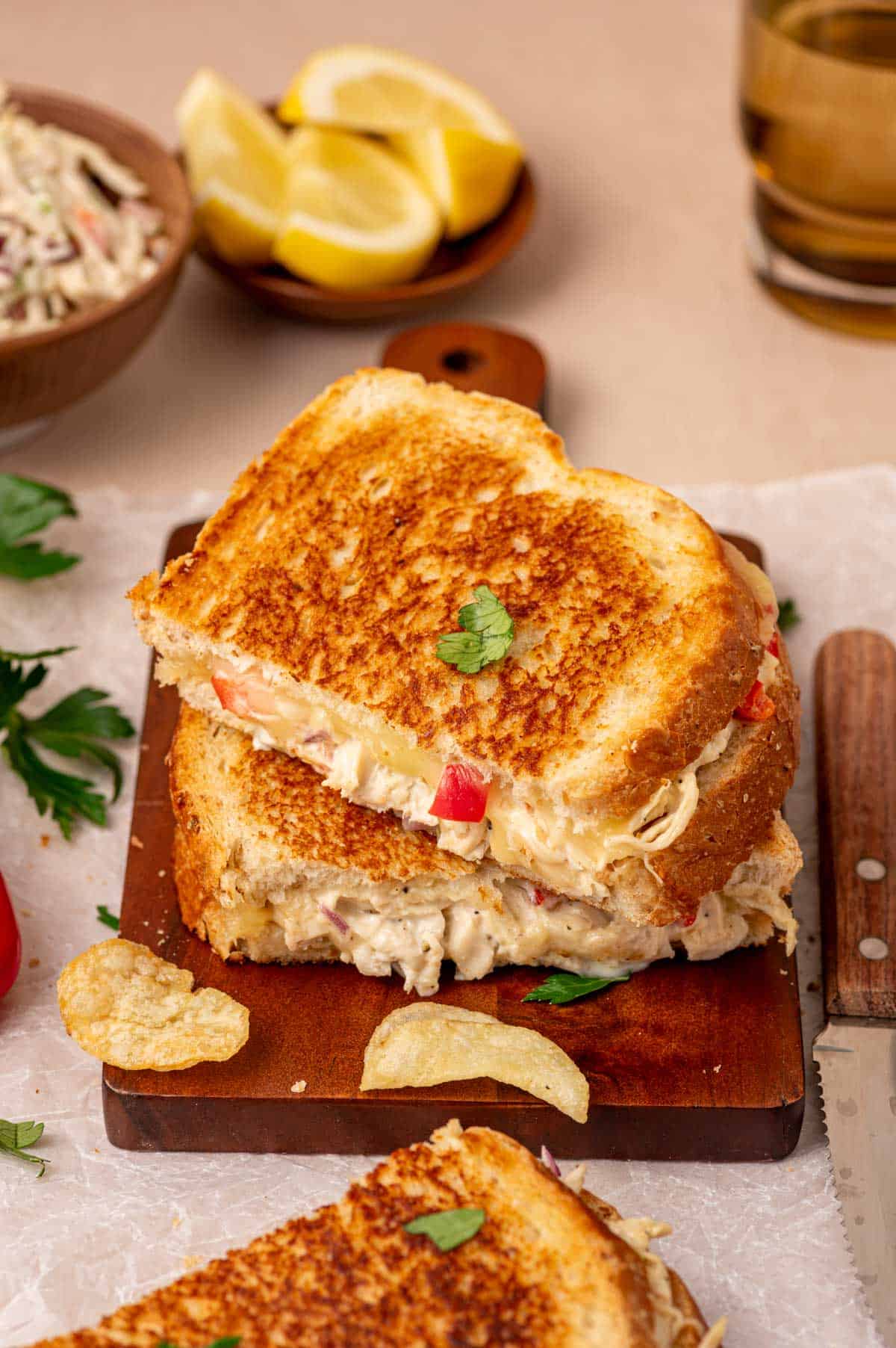 Warm and crispy chicken salad melt with gooey cheese, placed on a wooden board with a side of coleslaw  and Chips.